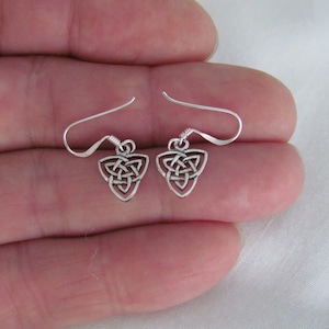 May include: A pair of silver Celtic knot earrings with a triangular shape. The earrings are on a hand.