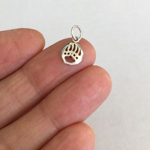 May include: A small, silver-colored charm featuring a bear paw print design. The charm has a loop at the top for attaching to a necklace or bracelet. The bear paw print is detailed with individual claw marks.