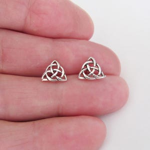 May include: A pair of small, silver-colored stud earrings. Each earring features a Celtic knot design, a symmetrical triangular shape with interwoven lines. The earrings are displayed on a light background.