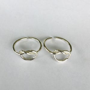 May include: Two silver-colored rings with a knot design. The rings are adjustable, with a small gap at the top. The rings are on a white background. The rings are likely made of sterling silver.