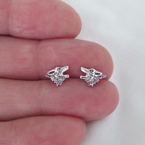 May include: A pair of small, silver-colored wolf head stud earrings. The wolf heads are detailed with open mouths and textured fur, set against a neutral background. These earrings are a unique accessory.