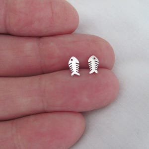 May include: A pair of small, silver-colored fishbone stud earrings. Each earring features a fish skeleton design with a rounded head and detailed rib-like structures. The earrings are displayed on a light background.