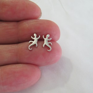 May include: A pair of silver-colored stud earrings. Each earring is shaped like a stylized lizard or gecko, with a small head, outstretched arms, and a curled tail. The earrings are small and delicate.