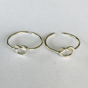 May include: Two silver-colored adjustable rings. Each ring features a knot design at the center. The rings are set against a plain, light-colored background. The rings are likely made of sterling silver or a similar metal.