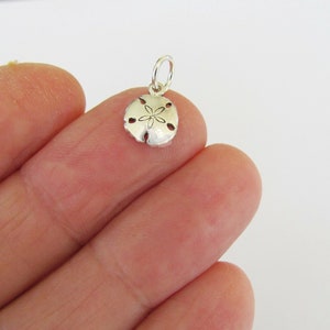 May include: A small, silver-colored sand dollar charm with a loop for attaching to a necklace or bracelet. The charm has a detailed design with a central flower-like pattern and several small holes around the edges. The charm is held between fingers.
