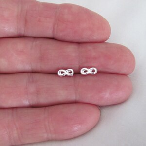 May include: A pair of small, silver-colored infinity symbol stud earrings. The earrings are displayed on a light background. The infinity symbol is a figure eight on its side, representing eternity and endless possibilities. These earrings are a simple and elegant accessory.