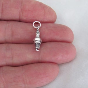 May include: A silver charm in the shape of a spark plug. The charm is small and detailed, with a realistic design.
