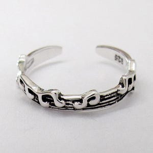May include: A silver-colored adjustable toe ring with an ornate, swirling design. The ring is open at the top and has a textured pattern along the band. The metal has a slightly antiqued appearance.