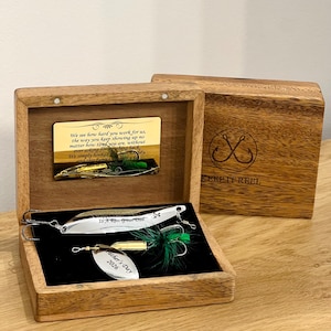 May include: A wooden box containing two silver and gold fishing lures with green feathers. The box lid features a gold plaque with text and a logo reading KEEP IT REEL. The lures are engraved with To A Reel Great Dad and Father's Day 2026.