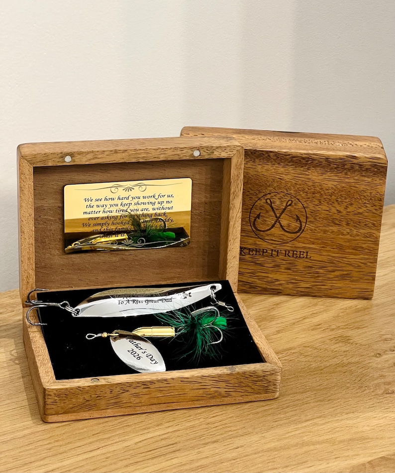 May include: A wooden box containing two silver and gold fishing lures with green feathers. The box lid features a gold plaque with text and a logo reading KEEP IT REEL. The lures are engraved with To A Reel Great Dad and Father's Day 2026.