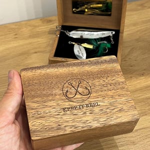 May include: A wooden box with a hinged lid featuring a laser-engraved circular fishing hook icon and the text KEEP IT REEL. Inside, a black velvet-lined compartment holds a silver-toned metal fishing lure with green feathers and a small tag.