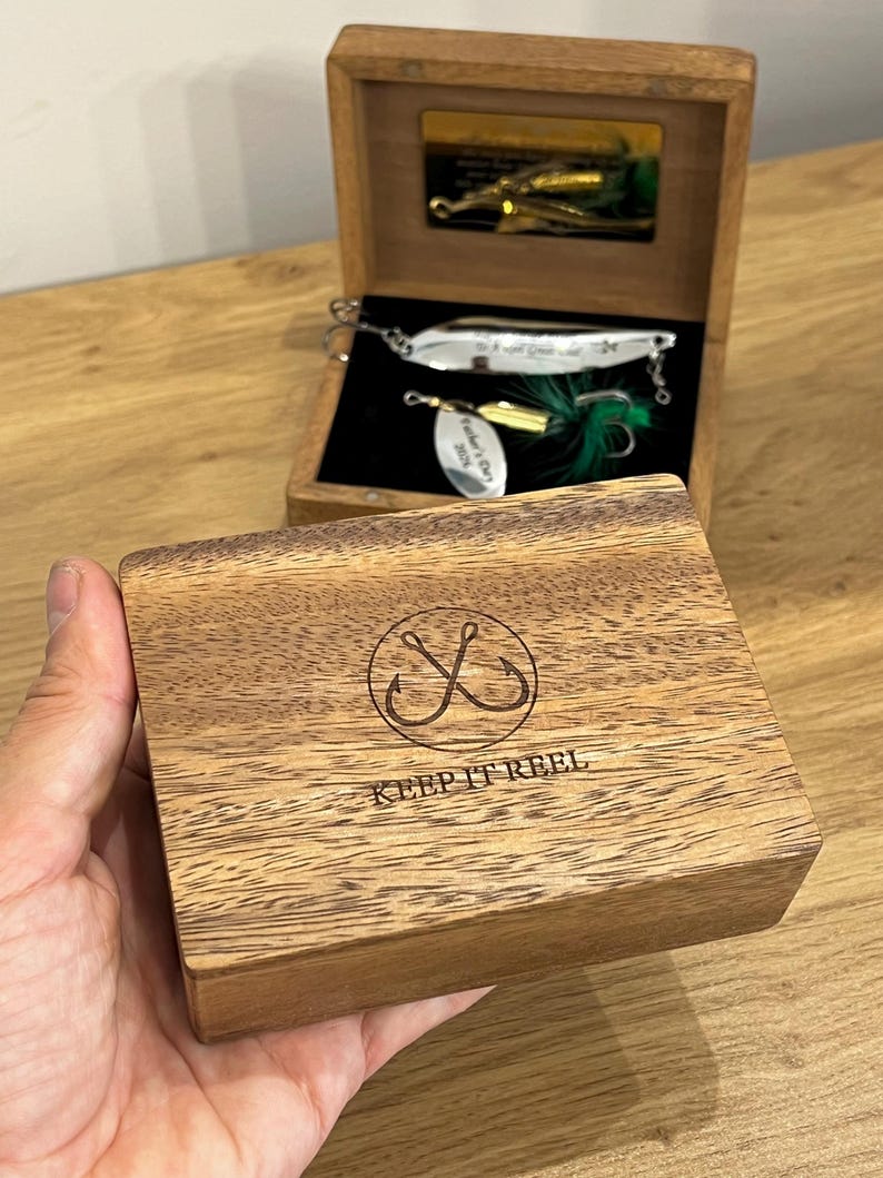 May include: A wooden box with a hinged lid featuring a laser-engraved circular fishing hook icon and the text KEEP IT REEL. Inside, a black velvet-lined compartment holds a silver-toned metal fishing lure with green feathers and a small tag.