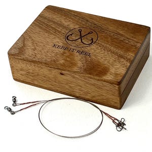 May include: A rectangular wooden box with a natural grain finish, featuring a circular logo with two crossed fishing hooks and the text "KEEP IT REEL" engraved on the lid. Beside the box lies a thin, dark metal fishing leader wire.