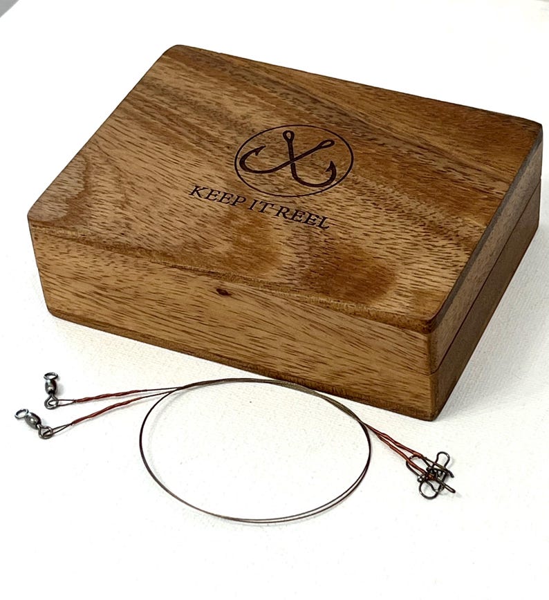 May include: A rectangular wooden box with a natural grain finish, featuring a circular logo with two crossed fishing hooks and the text "KEEP IT REEL" engraved on the lid. Beside the box lies a thin, dark metal fishing leader wire.