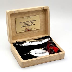 A bamboo box contains two fishing lures. One silver lure is inscribed with "O'Fish ally Retired." The other lure has the text "Cheers to the one that got away!" A small card reads "There are far better things ahead...Happy Retirement."