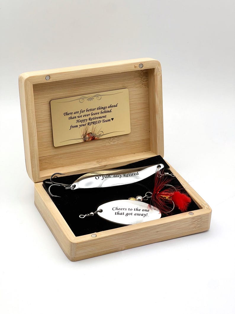 A bamboo box contains two fishing lures. One silver lure is inscribed with "O'Fish ally Retired." The other lure has the text "Cheers to the one that got away!" A small card reads "There are far better things ahead...Happy Retirement."