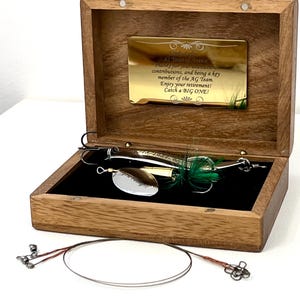 May include: A wooden box with a gold-toned plaque engraved with retirement text, containing a silver fishing lure with green feathers. The plaque reads: Cindy Eaf, 42 Years of Service, Thanks for your dedication, contributions, and being a key member of the AG Team. Enjoy your retirement! Catch a BIG ONE!