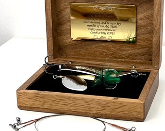 May include: A wooden box with a gold-toned plaque engraved with text thanking a team member for 42 years of service. Inside, a silver fishing lure with green feathers rests on a black velvet lining. A thin metal wire leader lies in front.