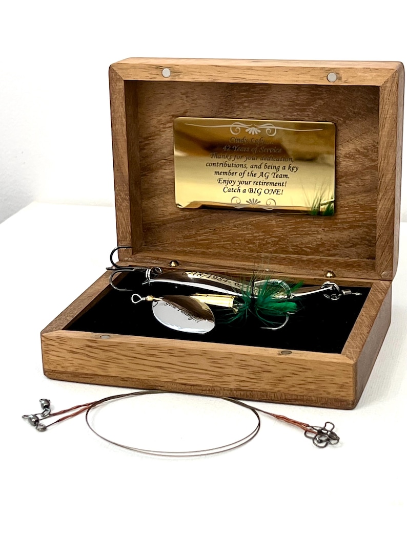 May include: A wooden box with a gold-toned plaque engraved with text thanking a team member for 42 years of service. Inside, a silver fishing lure with green feathers rests on a black velvet lining. A thin metal wire leader lies in front.