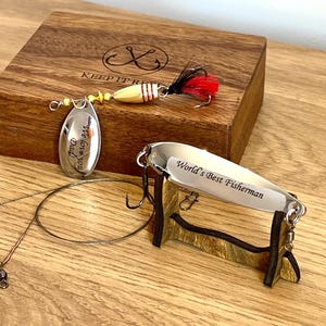 May include: Two metal fishing lures and a wooden box. One lure has a gold body, red and black feathers, and a silver blade engraved with "We love you Dad". The other is a silver spoon lure engraved with "World's Best Fisherman" on a stand.