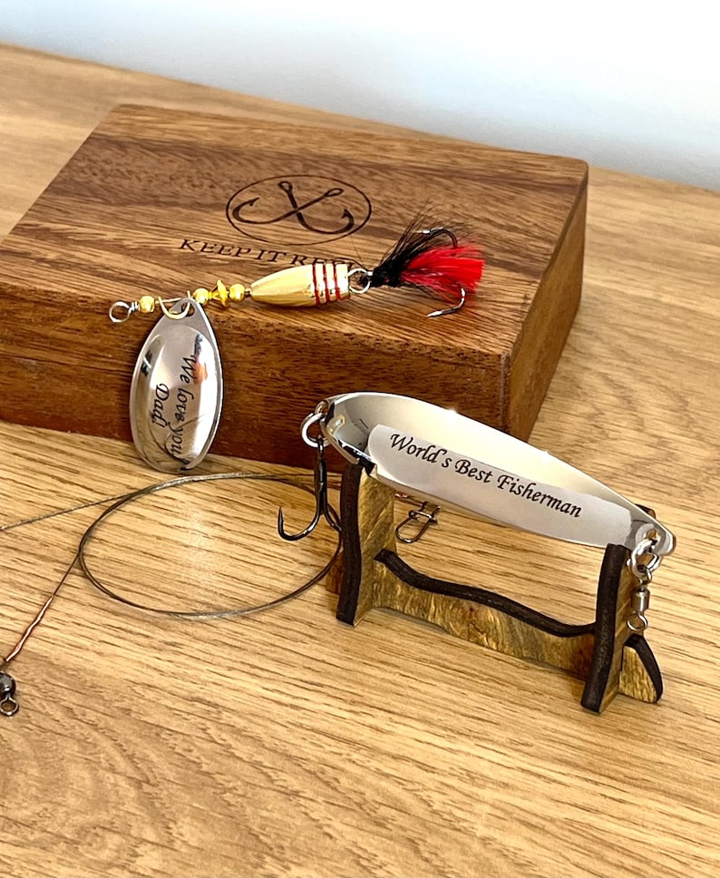 May include: Two metal fishing lures and a wooden box. One lure has a gold body, red and black feathers, and a silver blade engraved with "We love you Dad". The other is a silver spoon lure engraved with "World's Best Fisherman" on a stand.
