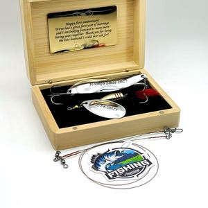 May include: A light-colored wooden box containing two silver fishing lures with hooks and red feathers. The box lid features a gold-toned metal plate with the text Happy first anniversary! We've had a great first year of marriage.