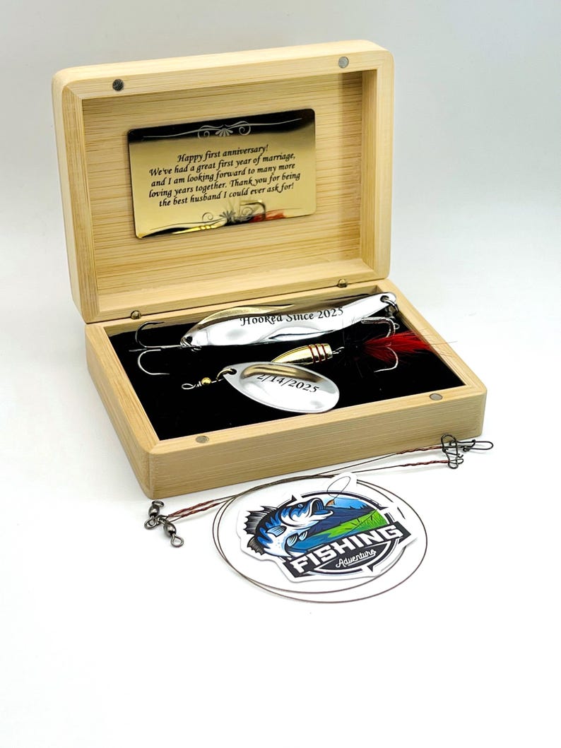 May include: A light-colored wooden box containing two silver fishing lures with hooks and red feathers. The box lid features a gold-toned metal plate with the text Happy first anniversary! We've had a great first year of marriage.