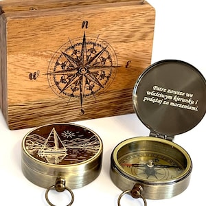 Peut inclure: Une boîte en bois avec une rose des vents gravée au laser et deux boussoles en laiton. Le couvercle de l'une présente un voilier, tandis que l'autre est ouverte, révélant un cadran et le texte polonais : Patrz zawsze we właściwym kierunku i podążaj za marzeniami.