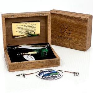 May include: A wooden box containing two metal fishing lures with green feathers and a silver tag engraved with "Father's Day 2026". A gold plaque inside the lid features a printed message. A circular "FISHING Adventure" tag lies in front.