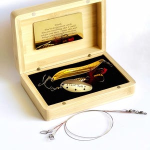 Op de afbeelding: Een open bamboe doos met twee vislokazen en een gouden kaart. De kaart leest "You're my greatest catch". Eén aas is goudkleurig met rode veren, de andere zilverkleurig met "I love you" gegraveerd.