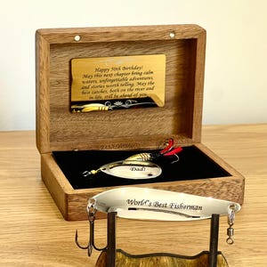 May include: A wooden box with a gold-toned plaque engraved with Happy 50th Birthday message. Inside, a fishing lure is engraved with Dad. A second silver-toned fishing lure engraved with World's Best Fisherman rests on a small wooden stand.