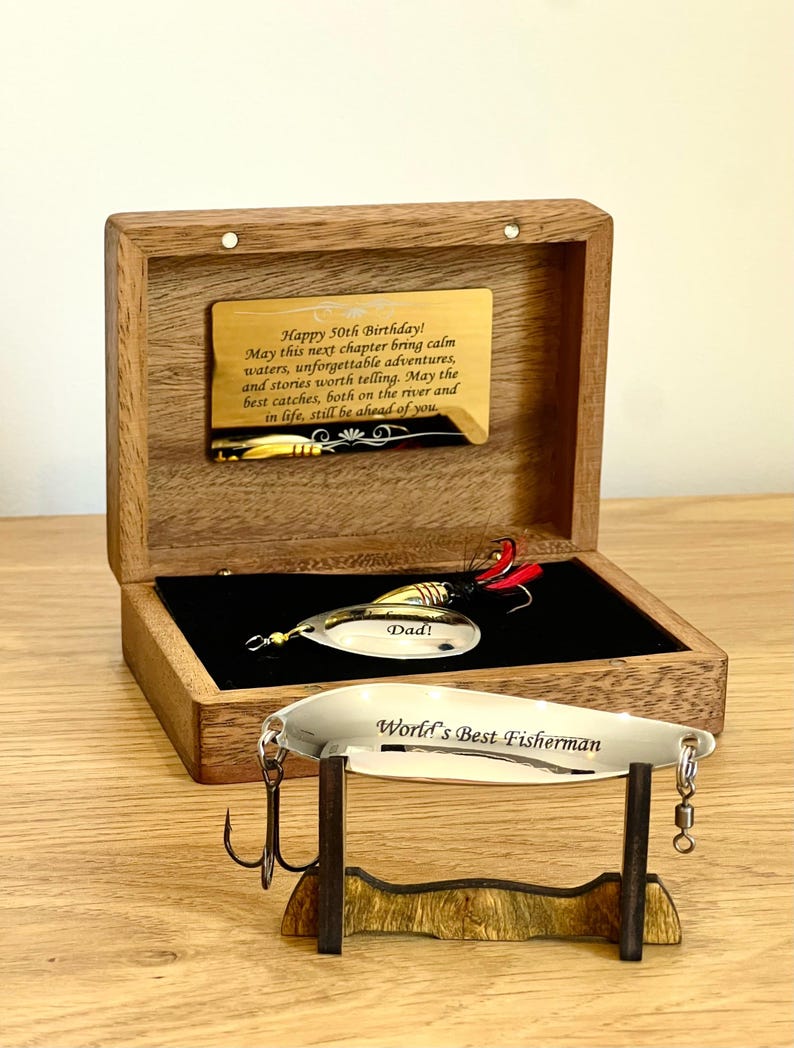 May include: A wooden box with a gold-toned plaque engraved with Happy 50th Birthday message. Inside, a fishing lure is engraved with Dad. A second silver-toned fishing lure engraved with World's Best Fisherman rests on a small wooden stand.