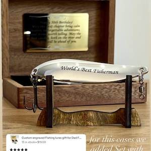 May include: A silver-colored metal fishing lure engraved with the text World's Best Fisherman, featuring a treble hook and swivel. The lure rests on a dark wooden stand in front of an open wooden box containing a gold-toned plaque with engraved text.