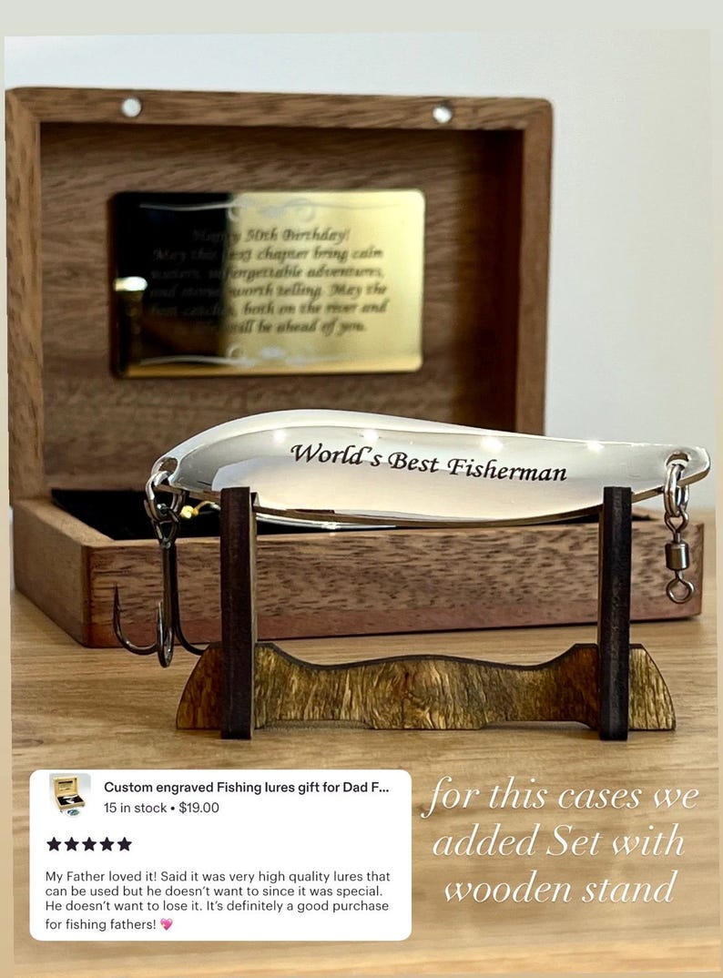 May include: A silver-colored metal fishing lure engraved with the text World's Best Fisherman, featuring a treble hook and swivel. The lure rests on a dark wooden stand in front of an open wooden box containing a gold-toned plaque with engraved text.