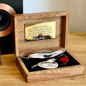 May include: A wooden box with a gold-toned metal plaque engraved with birthday wishes. Inside, two silver-colored fishing lures rest on black velvet. One lure is engraved with "World's Best Fisherman" and the other with "We love you, Dad!" featuring a red feather.
