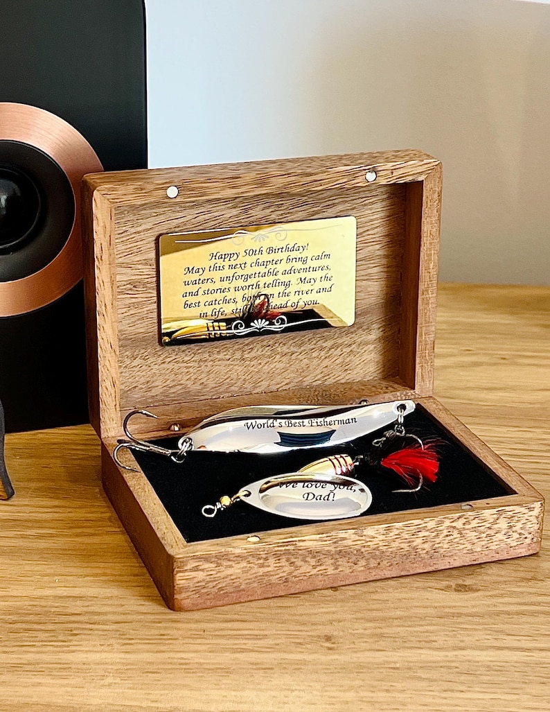 May include: A wooden box with a gold-toned metal plaque engraved with birthday wishes. Inside, two silver-colored fishing lures rest on black velvet. One lure is engraved with "World's Best Fisherman" and the other with "We love you, Dad!" featuring a red feather.
