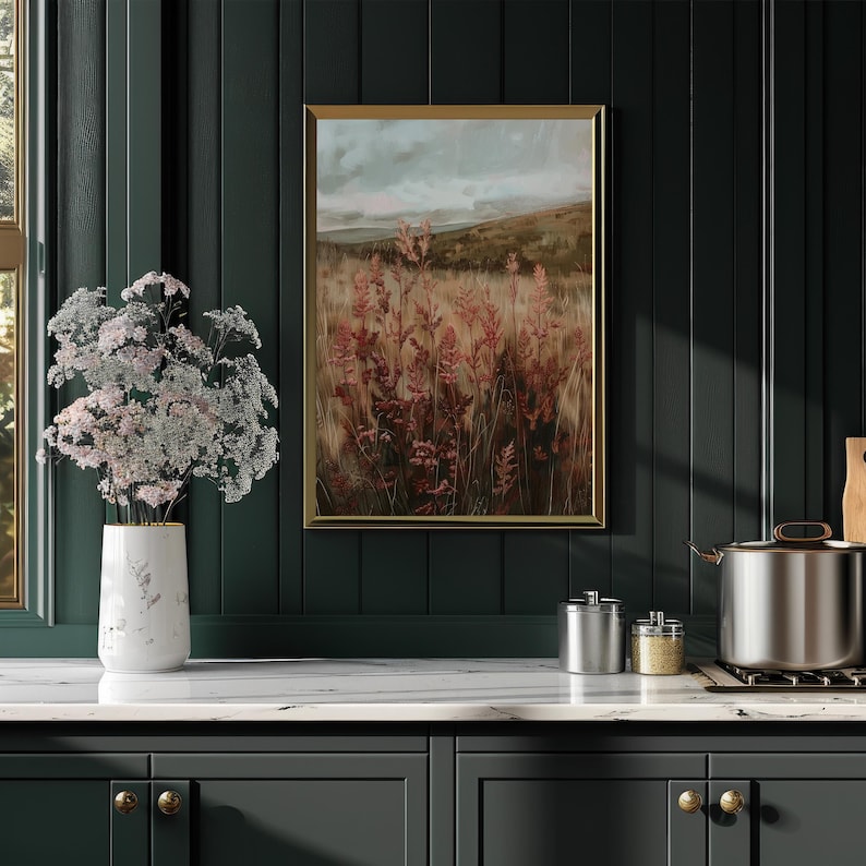 May include: A framed landscape painting with a gold frame depicts a field of tall, reddish-brown plants under a cloudy sky. A white vase with pink flowers sits on a white countertop. The background is a dark green wall.
