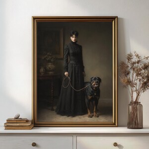May include: A framed portrait of a woman in a long black dress, holding a leash attached to a Rottweiler dog. The painting has a gold frame and is displayed on a white surface. The dog is black and brown, and the woman is standing in a room with a table and vase.