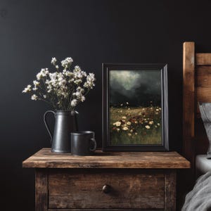 May include: A still life arrangement on a wooden nightstand. It features a black framed painting of a field of flowers, a black pitcher with white flowers, and a black mug. The background is a dark wall.