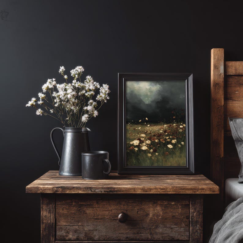 May include: A still life arrangement on a wooden nightstand. It features a black framed painting of a field of flowers, a black pitcher with white flowers, and a black mug. The background is a dark wall.