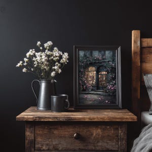 May include: A still life featuring a dark metal pitcher with white flowers, a black mug, and a framed picture of a building with a floral facade. The items are on a wooden nightstand next to a bed.
