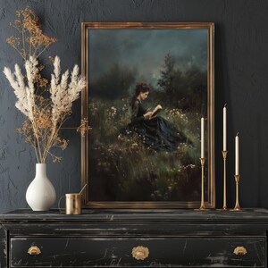 May include: A framed oil painting of a woman in a black dress sitting in a field of wildflowers, reading a book. The painting has a dark, moody atmosphere and is set against a dark gray background.