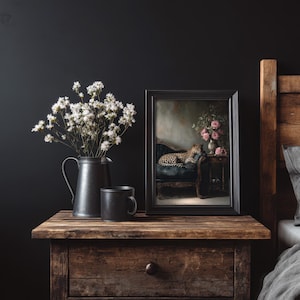 May include: A dark, moody still life. A framed picture of a leopard on a blue velvet chair sits on a wooden bedside table, alongside a black jug of white flowers and a black mug. The scene is set against a dark wall, creating a dramatic effect.