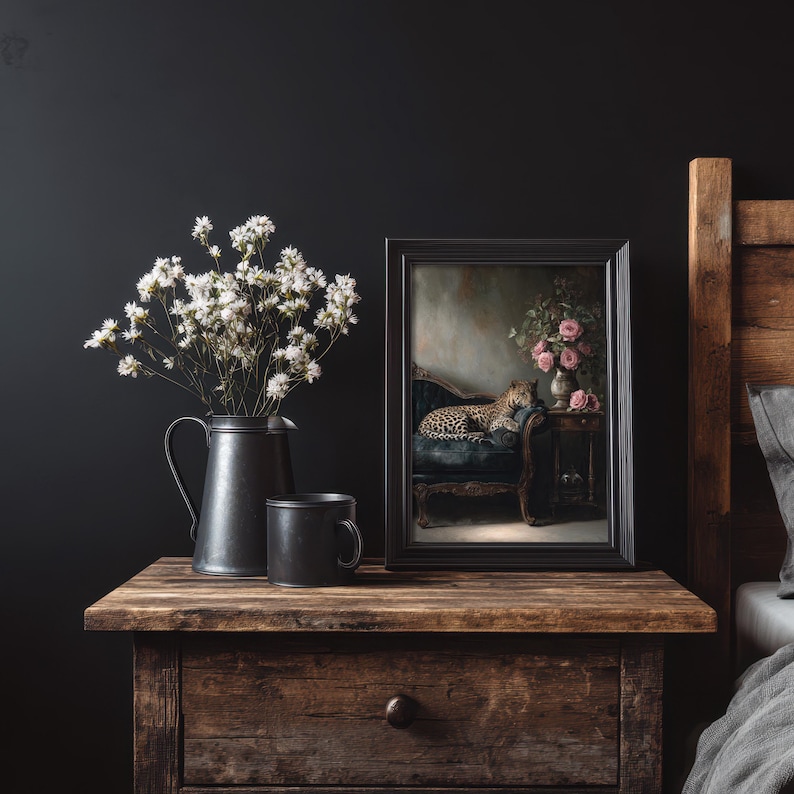 May include: A dark, moody still life. A framed picture of a leopard on a blue velvet chair sits on a wooden bedside table, alongside a black jug of white flowers and a black mug. The scene is set against a dark wall, creating a dramatic effect.