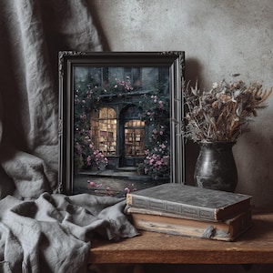 May include: A framed artwork depicts a quaint bookstore facade, adorned with climbing roses. Two antique books rest on a wooden surface, alongside a vase of dried flowers. A gray cloth drapes in the foreground, creating a vintage aesthetic.