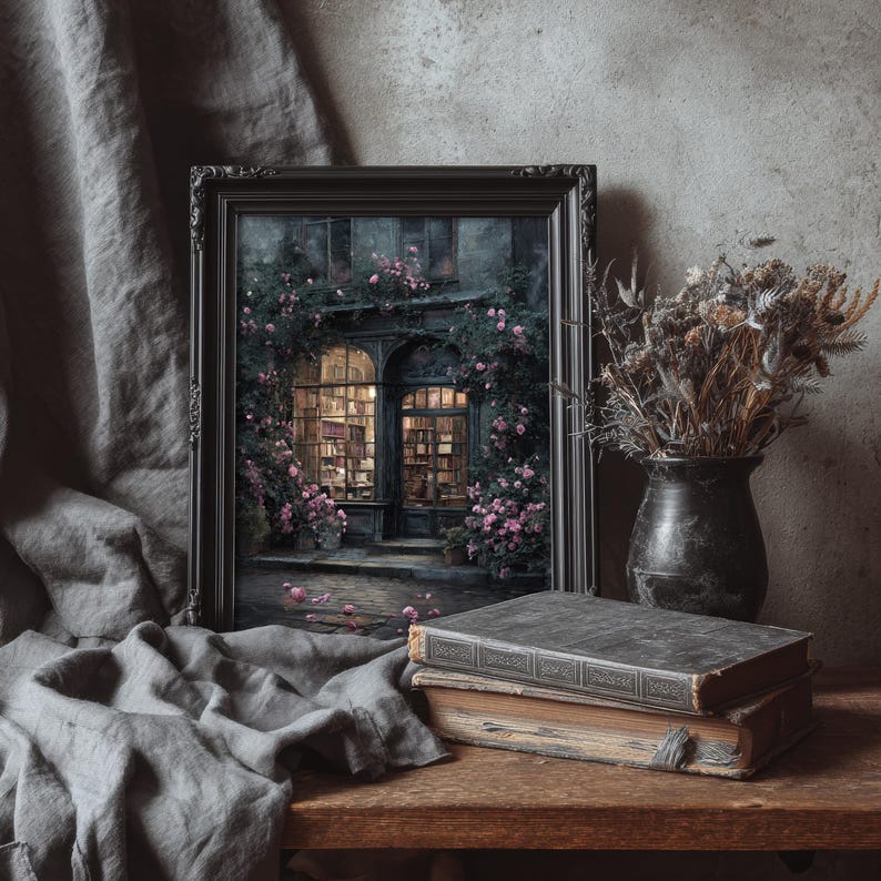 May include: A framed artwork depicts a quaint bookstore facade, adorned with climbing roses. Two antique books rest on a wooden surface, alongside a vase of dried flowers. A gray cloth drapes in the foreground, creating a vintage aesthetic.