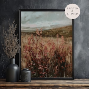 May include: Framed landscape art print featuring a field of tall, reddish-brown plants in the foreground, with a backdrop of rolling hills and a cloudy sky. The artwork is in a black frame and displayed on a wooden surface.