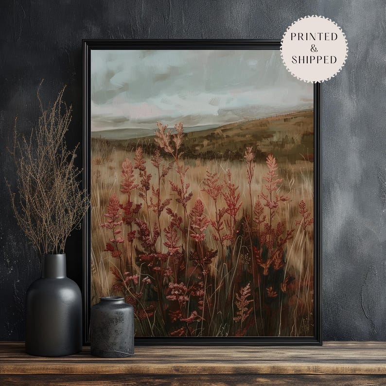May include: Framed landscape art print featuring a field of tall, reddish-brown plants in the foreground, with a backdrop of rolling hills and a cloudy sky. The artwork is in a black frame and displayed on a wooden surface.