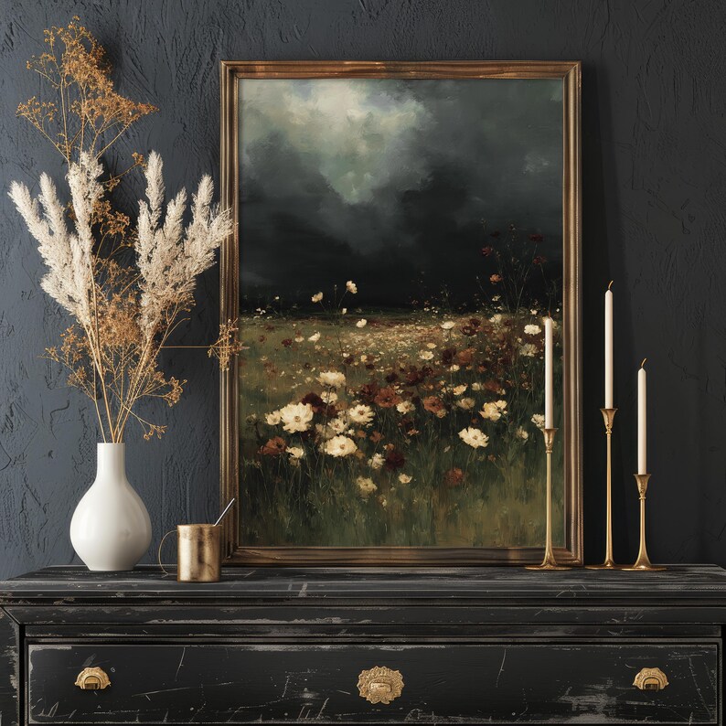 May include: A framed landscape painting depicts a field of white and red flowers under a dark, stormy sky. The painting is displayed on a black dresser, alongside a white vase with dried flowers, a gold mug, and brass candle holders.