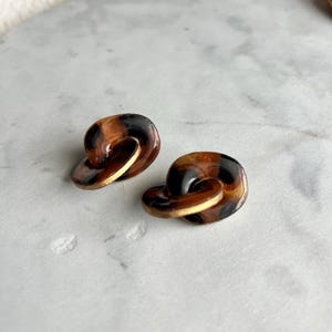 May include: A pair of vintage-style earrings. Each earring has a layered, circular design with a tortoiseshell pattern in brown and black. The earrings have a gold-coloured base and are displayed on a marble surface.